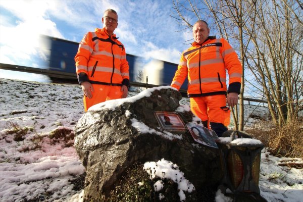 Ralf Kowalleck (rechts) und Andreas Stratmann stehen an einem Stein, der an einen Kollegen der beiden Straßenmeister erinnern soll. 2001 verstarb er wenige Meter hinter den beiden auf der A44, als ihn ein Auto in einem abgesperrten Bereich erfasste. - © Lukas Brekenkamp
