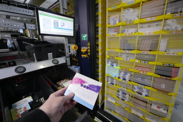 Beim Picken: Der Computer zeigt LZ-Redakteur Jost Wolf auf dem Monitor, dass nun die Plaque-Färbetabletten aus Fach 1E entnommen werden müssen. Zusätzlich wird das Fach quadratisch hell angestrahlt. Das gelbe Regal hat vorher ein Roboter an die Öffnung der Pick-Station gefahren. Die Ware muss nach dem Scannen in eine der schwarzen Kisten gelegt werden. Das wird mit Druck auf den runden schwarzen Knopf darüber bestätigt. - © Jost Wolf