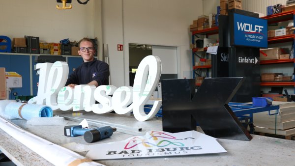 Azubi Lukas Hannemann steht hinter dem Schriftzug der Bäckerei Hensel – der wird noch beleuchtet und bekommt Farbe. Bei „Infographik“ werden auch touristische Schilder, Buchstaben sowie Werbepylone wie im Hintergrund gefertigt. - © Alexandra Schaller