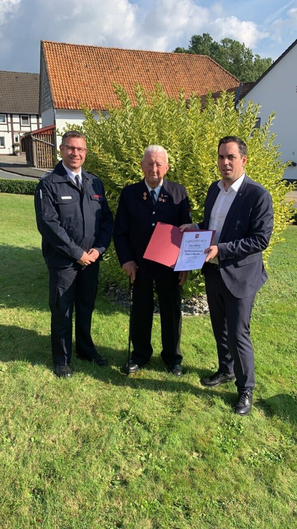 Der Leiter der Feuerwehr Lügde, Michael Schoppmeier (links), und Bürgermeister Torben Blome nehmen August Moseke in ihre Mitte.  - © Feuerwehr Lügde