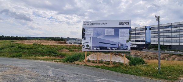 Auf dem Bauplakat zeigt sich bereits, wie es mal aussehen soll, und im Hintergrund haben die Bagger bereits eine Fläche für das neue Logistikzentrum freigeräumt, wo vor einigen Wochen noch ein Hügel war. - © Marianne Schwarzer