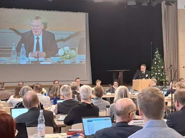Per Livestream zu den Mitgliedern: Während Landessuperintendent Dietmar Arends - groß auf der Leinwand zu sehen - zu den Synodalen spricht, können Interessierte die Sitzung online verfolgen. Am Rednerpult steht Präses Michael Keil. - © Carolin Brokmann-Förster