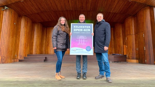 Leonie Immel (von links, Eventmanagement), Bürgermeister Dirk Tolkemitt und Oliver Siekmann (Stadtmarketing) präsentieren das Plakat zum Silvester Open-Air in der Konzertmuschel im Kurpark. - © Stadt Bad Salzuflen