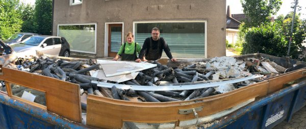 Nathalie Witt und Max König stehen vor einer von vier großen Mulden mit Abfall aus dem vollgelaufenen Keller ihres Hauses (im Hintergrund). - © Thomas Reineke
