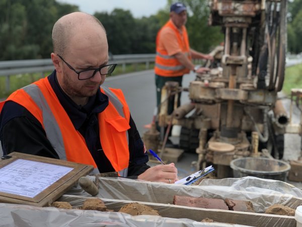 Projektgeologe Niko Miera macht eine erste Sichtungsprobe des Gesteins, auf dem die neue B 66 zwischen Barntrup und Großenmarpe einst verlaufen soll. Im Hintergrund holt der Kollege mit dem Bohrgerät Meter um Meter aus dem Untergrund hervor. - © Till Brand