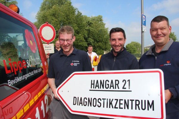 Domenik Sonntag (Deutsches Rotes Kreuz), Dennis Kuhlmann und Christoph Bergmann (beide Eigenbetrieb Straßen) schrauben ein Schild am Hangar 21 ab. Hinten: Jochen Steinkühler (Bevölkerungsschutz; links) und Jason Jochem (Deutsches Rotes Kreuz). - © Martin Hostert(LZ)