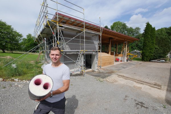 Nick Blandau zeigt ein Stück der Fernwärme-Leitung, von der er mit Familie, Freunden und Nachbarn zwei Kilometer in Vahlhausen in der Erde verlegt hat. Im Hintergrund ist der Schuppen mit der Holzhackschnitzelheizung zu sehen, auf dessen Dach gerade eine Fotovoltaikanlage installiert wird. - © Jost Wolf