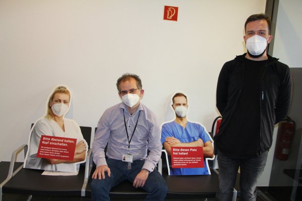 Pappaufsteller, für die Krankenhaus-Mitarbeiter vor der Kamera gestanden haben, dienen im Klinikum als Abstandshalter. Chefarzt Daniel Fischer (sitzend) und Martin Wall von der Klinik-Einsatzleitung werben dafür, die Kontakte einzuschränken. - © Astrid Sewing