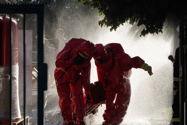 Feuerwehrleute in chemischen Schutzanzügen retten einen Verletzten, während im Hintergrund die giftigen Gase mit Wasser niedergeschlagen werden. - © Freitag-TV