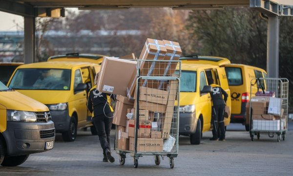 Eine Mitarbeiterin der Deutsche Post DHL zieht einen vollen Paketwagen zu ihren Fahrzeug über den Hof eines Zustellpunktes. - © Jan Woitas/dpa/Archivbild