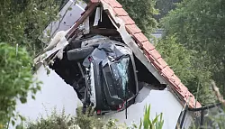 Das Auto liegt nach einem kurzen Flug seitlich in der Mauer einer Scheune. - © -/Moonshine Media/dpa