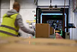 Ein DHL-Mitarbeiter nimmt Pakete in Emfpang - im Hintergrund ist ein Roboter zu sehen, der die Pakete aus einem Container nimmt und sie auf ein F&ouml;rderband legt. (Archivbild) - &copy; Wolf von Dewitz/dpa