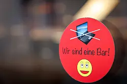 An einer Scheibe der Café-Bar 8tto Grammi in Bayreuth weist ein Schild auf das Verbot von Laptops hin. - © Daniel Karmann/dpa