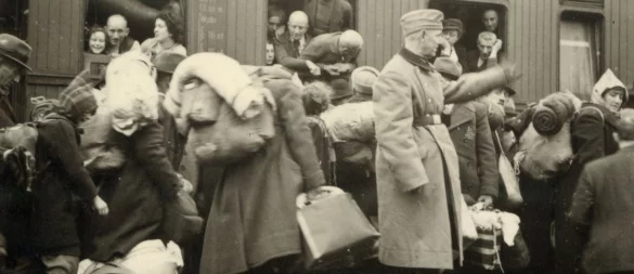 Jüdinnen und Juden am Bielefelder Hauptbahnhof auf dem Weg zu den ihnen zugewiesenen Waggons. Im Zug sitzen bereits Deportierte aus dem Kreis Münster. Im Vordergrund steht ein Mitglied des Begleitkommandos mit erhobener Hand. - © Stadtarchiv Bielefeld