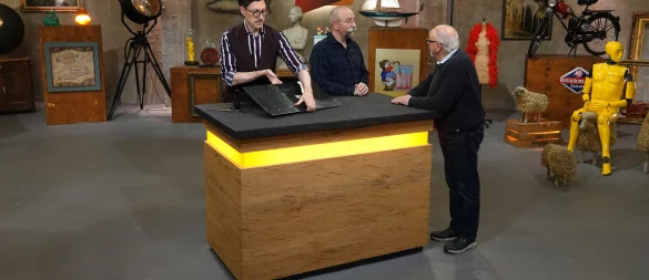 Experte Colmar Schulte-Goltz (v.l.) begutachtet bei "Bares für Rares" das Miele-Emailleschild, während Moderator Horst Lichter mit dem Harsewinkeler Georg Terlutter redet. - © ZDF/Warner