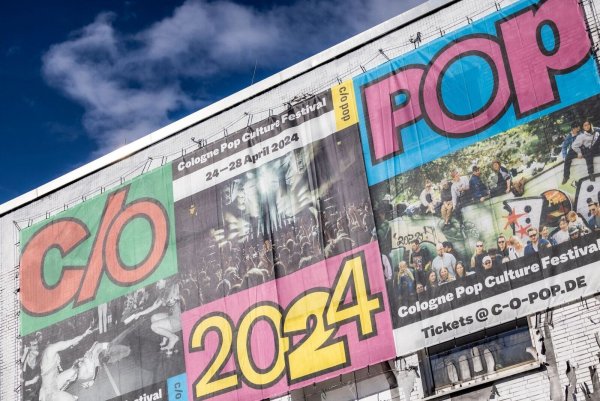 An einem Gebäude im Stadtteil Ehrenfeld hängt ein großes Banner, dass für das Musikfestival "c/o pop" wirbt. - © Thomas Banneyer/dpa