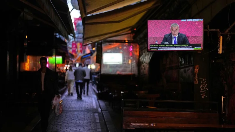 Menschen gehen an einem Fernsehbildschirm vorbei, auf dem der Pr&auml;sident des T&uuml;rkischen Fu&szlig;ballverbands, Ibrahim Haciosmanoglu, w&auml;hrend einer Pressekonferenz zu sehen ist. - &copy; Francisco Seco/AP/dpa