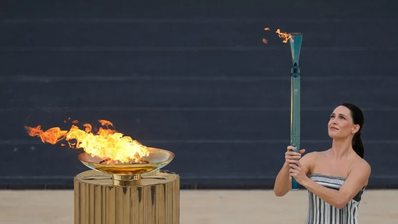 Schauspielerin Mary Mina, in der Rolle der Hohepriesterin, entzündet eine Fackel bei der Zeremonie zur Übergabe der Flamme der Olympischen Winterspiele 2026 im Panathinaikon-Stadion. - © Petros Giannakouris/AP/dpa