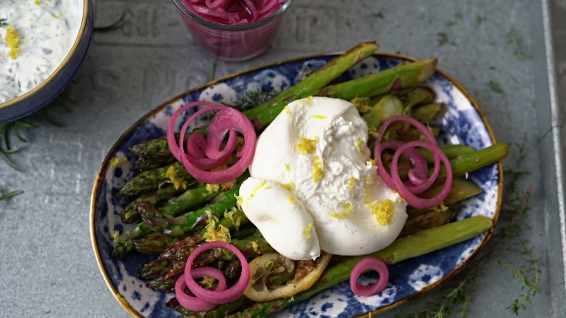 Zum Anrichten wird der Burrata über den Spargelstangen etwas auseinander gezupft und mit dem Ranchdip beträufelt. Zuvor eingelegte rote Zwiebelringe bringen Farbe auf den Teller. - © Mareike Pucka/biskuitwerkstatt.de/dpa-tmn