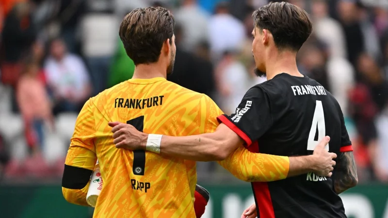 Kevin Trapp (l) fehlt im Pokal-Kader von Eintracht Frankfurt und hat die Kapitänsbinde an Robin Koch abgegeben. - © Jan-Philipp Strobel/dpa