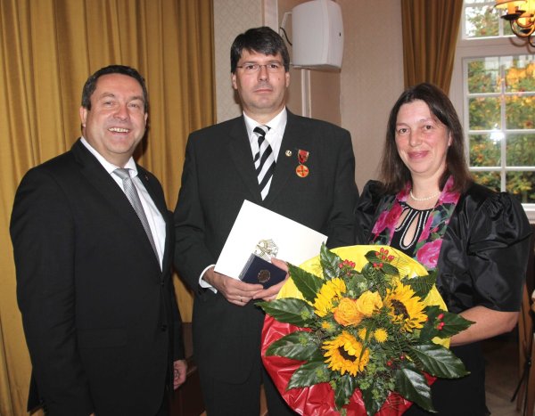 Ausgezeichnet: Stephan Grigat (Mitte) erhält die Verdienstmedaille von Landrat Axel Lehmann, seine Frau Julia bekommt Blumen. - © privat
