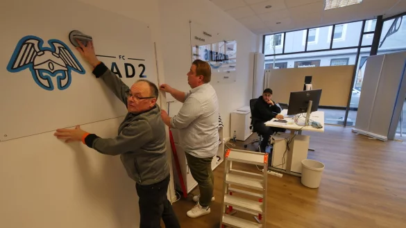 Rene P&auml;tzig und Pressesprecher Oliver Kenschke bringen das Logo des Amazon-Logistikzentrums in Belle an. FC steht f&uuml;r "Fulfilment Center" und PAD2 ist das K&uuml;rzel des n&auml;chsten Flughafens Paderborn. PAD1 steht in Oelde. Das Logo links ist vom Hermannsdenkmal inspiriert. Im Hintergrund wartet Shota Kurtsikidze auf Job-Interessenten. - &copy; Jost Wolf