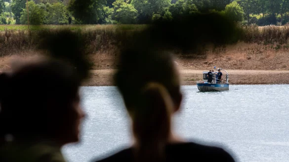 Personen blicken auf ein Polizeiboot, das &uuml;ber den Kiesteich f&auml;hrt, an dem am Sonntag aus ungekl&auml;rten Gr&uuml;nden beim Baden zwei Personen nicht mehr aufgetaucht sind. - &copy; Lino Mirgeler/dpa