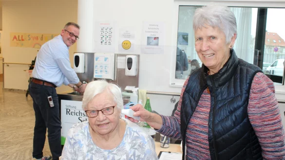 Gisela Langer (rechts) geh&ouml;rt zu den Ehrenamtlichen, die das Screening an der Pforte &uuml;bernehmen und zeigt mit Bewohnerin Gerda Salobir, dass bei jedem Besucher erst Fieber gemessen und dann ein Schnelltest durchgef&uuml;hrt wird. Einrichtungsleiter Harm-Hendrik M&ouml;ller (im Hintergrund) ist dankbar f&uuml;r die Unterst&uuml;tzung. - &copy; Sandra Castrup