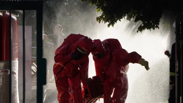 Feuerwehrleute in chemischen Schutzanz&uuml;gen retten einen Verletzten, w&auml;hrend im Hintergrund die giftigen Gase mit Wasser niedergeschlagen werden. - &copy; Freitag-TV