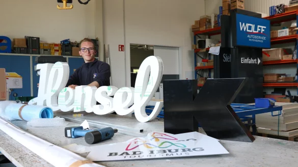 Azubi Lukas Hannemann steht hinter dem Schriftzug der Bäckerei Hensel – der wird noch beleuchtet und bekommt Farbe. Bei „Infographik“ werden auch touristische Schilder, Buchstaben sowie Werbepylone wie im Hintergrund gefertigt. - © Alexandra Schaller