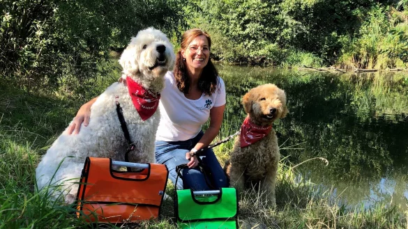 Katja Eggers sitzt mit &bdquo;Marlo" (links) und &bdquo;Bruno" am Ufer der Bega in den Hoffmannswiesen. Mit den Taschen, k&ouml;nnen die Hunde Therapiespielzeug transportieren.&nbsp; - &copy; Sven Kienscherf