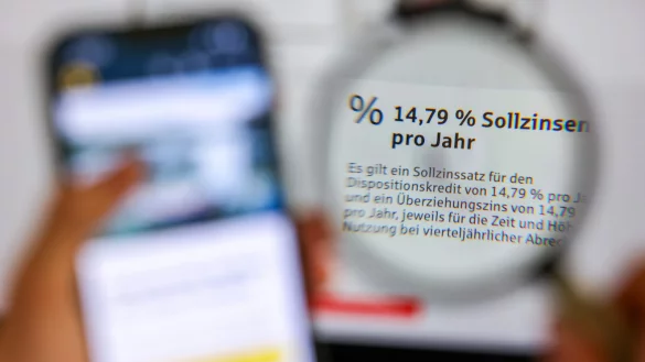 Durch eine Lupe kann man den Schriftzug „14,79% Sollzinsen pro Jahr“ für einen Dispositionskredit bei einer Sparkasse erkennen. Solche Zinshöhen werden auch in OWL schon erreicht und übertroffen. - © picture alliance/dpa