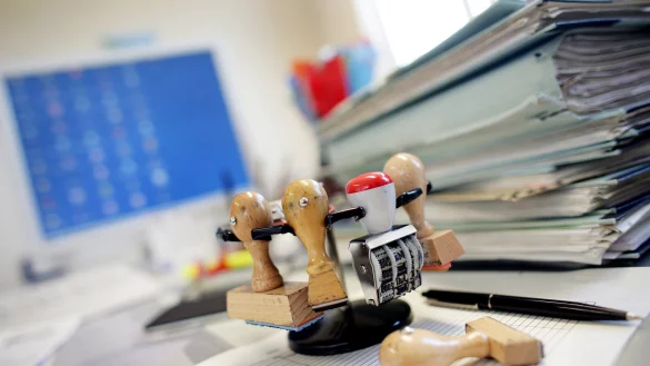 Ein Stempelhalter steht neben einem Stapel Akten auf einem Schreibtisch in einer Behörde. F. stellt sich immer häufiger die Frage, ob er als Beamter in einer Landesbehörde in OWL langfristig glücklich sein kann. - © Patrick Pleul/dpa (Symbolfoto)