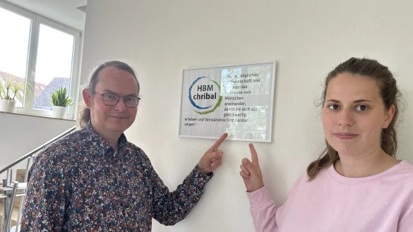 Pfarrer Mathias Zizelmann (links) und Linda Brandau zeigen auf das Motto von HMB "chribal". "Wir erm&ouml;glichen Gemeinschaft und wecken das Interesse der Menschen aneinander, damit sie sich als gleichwertig erleben und Verst&auml;ndnis f&uuml;reinander zeigen." - &copy; Karlotta Hensch