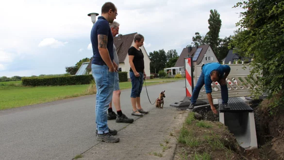 Sven Anders (rechts) zeigt, wo die Grabenkante abgesichert wird. Sein Kollege Sven Ranft (links) beantwortet ebenfalls die Fragen der Anwohner Marco Wiebrok und Katja Brokerhoff. Deren Hund &bdquo;Fina" l&auml;uft nicht gerne &uuml;ber die dicke Rinne. - &copy; Astrid Sewing