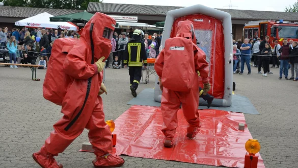 Zwei Feuerwehrleute haben mit Hilfe zweier Kameraden die roten, schweren Chemikalienschutzanz&uuml;ge &uuml;bergezogen. Hier zeigen beide, dass es nach einem Einsatz notwendig ist, in einer Spezialdusche die giftigen Stoffe abzuwaschen. - &copy; Karin Prignitz