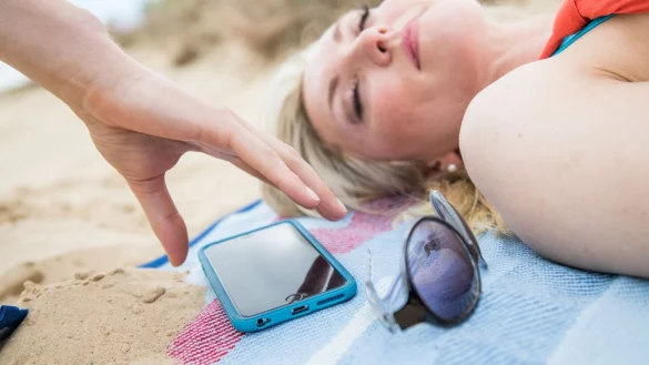 Eine Hand greift am Strand nach dem Handy einer sich Sonnenden. - &copy; Foto: Christin Klose/dpa-tmn