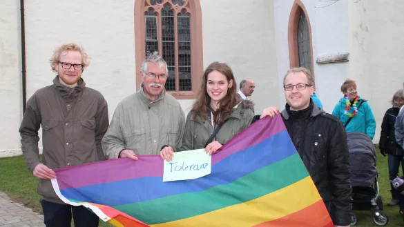 Rund 100 Gläubige wie Manfred Rothermel (2. v. l.) und Johanna Meier waren mit Regenbogenflagge dem Aufruf zum stillen Protest von Joachim Prüßner (links) und seinem Cousin Dominic (rechts) am Sonntag vor die Pauluskirche in Hohenhausen gefolgt. Im Hintergrund stellt sich Pfarrer Peter Busse der Menge. - © Judith Stracke