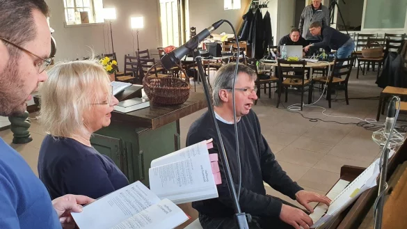 Vikar Gregor Block (von links), Pastorin Bettina Hanke-Postma und Superintendent Holger Postma proben f&uuml;r den ersten Online-Gottesdienst. Im Hintergrund ist das Technik-Team um Social-Media-Pfarrer Wolfgang Loest mit der Technik besch&auml;ftigt, die zwei Tische beansprucht.&nbsp; - &copy; Uwe Rotkamp