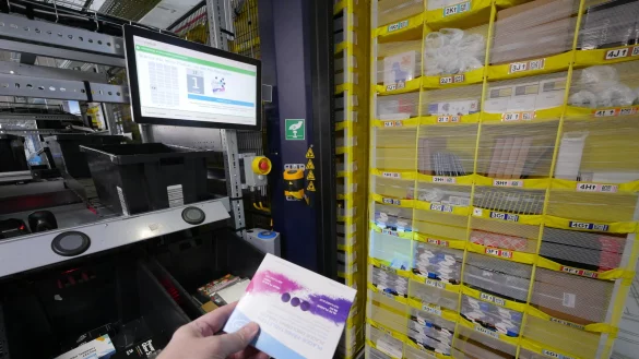 Beim Picken: Der Computer zeigt LZ-Redakteur Jost Wolf auf dem Monitor, dass nun die Plaque-F&auml;rbetabletten aus Fach 1E entnommen werden m&uuml;ssen. Zus&auml;tzlich wird das Fach quadratisch hell angestrahlt. Das gelbe Regal hat vorher ein Roboter an die &Ouml;ffnung der Pick-Station gefahren. Die Ware muss nach dem Scannen in eine der schwarzen Kisten gelegt werden. Das wird mit Druck auf den runden schwarzen Knopf dar&uuml;ber best&auml;tigt. - &copy; Jost Wolf