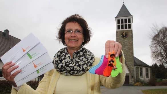 Voller Tatendrang: Pfarrerin Iris Beverung hält die Briefumschläge mit den neuen Logos für die Gemeinden Cappel und Istrup in ihrer rechten Hand. In ihrer linken zeigt sie kleine Glöckchen und Söckchen, die für die Fundraising-Aktion gedacht sind. Unter anderem soll damit Geld für eine neue Heizung für die Kirche in Istrup (im Hintergrund) gesammelt werden. - © Patrick Bockwinkel