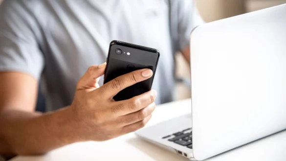 Mann mit Handy in der Hand vor einem Laptop - &copy; Foto: Zacharie Scheurer/dpa-tmn