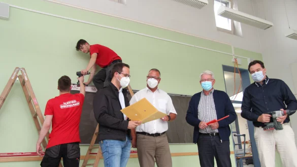 Joel Ollermann (von links) und Luca Klausing von der Firma Elektro Dresselhaus schrauben Kabelkan&auml;le an die W&auml;nde eines Klassenzimmers, w&auml;hrend Bauamtsleiter Manuel Br&ouml;ker, Maik Czarnecki aus dem Fachbereich Bildung, Soziales und Sport, B&uuml;rgermeister Thomas Katzer und K&auml;mmerer Patrick Herrmann einen Blick in die Pl&auml;ne werfen. - &copy; Nadine Uphoff