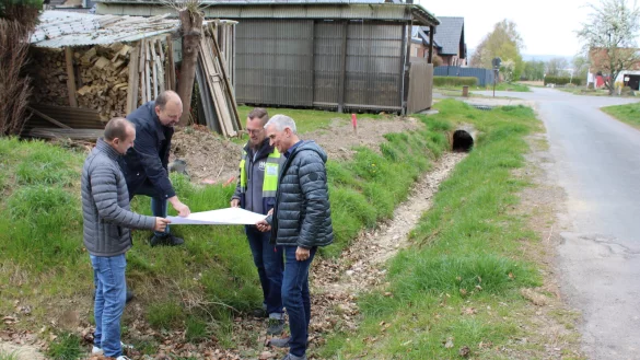 Abwasseringenieur Sven Ranft (von links), Sven Anders (Fachteamleiter Stadtentwässerung, Wasserwirtschaft), Tim Nehm (Westfalen Weser) und Beigeordneter Thorsten Paulussen schauen sich vor Ort die Pläne an. Im Hintergrund ist der Ablauf zu sehen, der nun vergrößert werden soll. Ebenso die Bodenschwelle, die versetzt wird. - © Lorraine Brinkmann