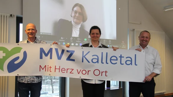 Die neuen Ärzte für Kalletal, Frank Norrenbrock (von links) und Imola Kalló, halten zusammen mit Bürgermeister Mario Hecker ein Banner mit dem Logo des MVZ. Im Hintergrund ist Geschäftsführerin Gabriele Dostal zu sehen, die online zugeschaltet ist. - © Nadine Uphoff