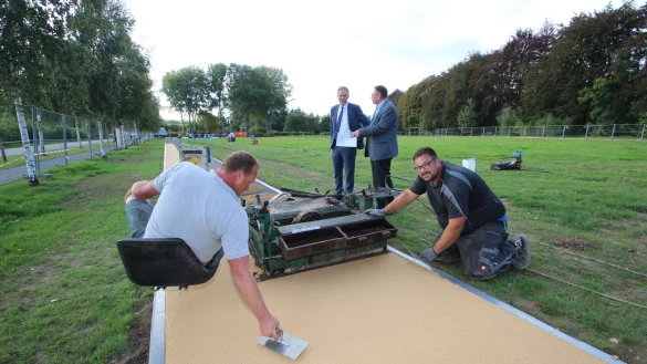 Alexander Lange (rechts) und Andr&eacute; Uhl tragen die Deckschicht f&uuml;r die Laufbahn auf, w&auml;hrend sich im Hintergrund Bauamtsleiter Steffen Schenk (rechts) und B&uuml;rgermeister J&uuml;rgen Schell auf der Faulen Wiese unterhalten. - &copy; Jens Rademacher