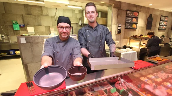 Raphael Schwuchow, Abteilungsleiter der Frischtheke im Bad Meinberger Edeka Wiele (links), und sein Kollege Josia Leischner vom Barntruper Marktkauf zeigen die Mehrwegbehälter, in denen Kunden Gerichte vom warmen Mittagstisch mitnehmen können, und das Blech, auf das mitgebrachte Behältnisse für Käse und Wurst gestellt werden. - © Jens Rademacher