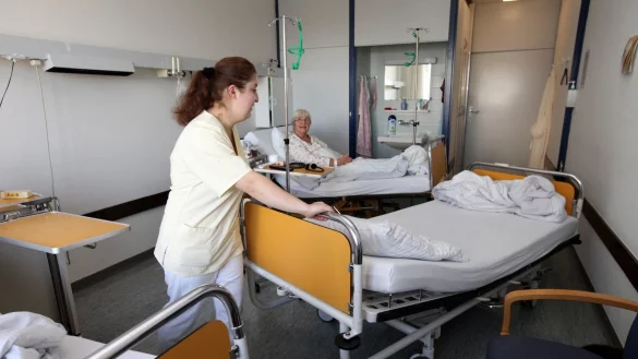 Eine Kliniksericekraft schiebt ein Bett aus einem Patientenzimmer auf der Station 3a. Im Hintergrund ist die Waschnische zu sehen, die sich die Zimmergemeinschaft teilen muss. Die Toilette ist auf dem Flur. - &copy; Bernhard Preu&szlig;