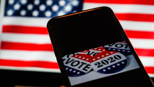 Abzeichen f&uuml;r die Pr&auml;sidentschaftswahl mit den Aufschriften &laquo;Vote&raquo; und &laquo;2020&raquo; sind auf einem Smartphone-Bildschirm vor einer US-Amerikanischen Flagge zu sehen. - &copy; (c) ZUMA Wire
