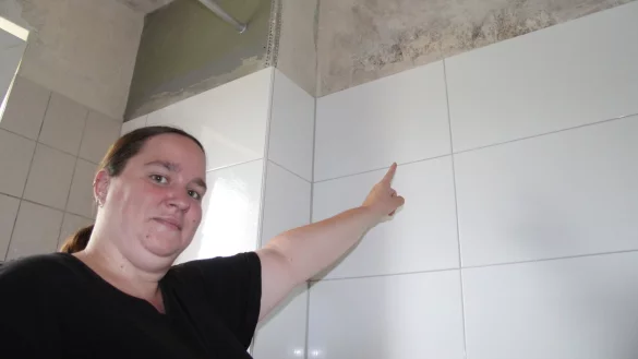 Katharina Korth weist auf die Stelle der Badezimmerwand, wo der vom Wasserschaden verursachte Schimmel immer noch zu sehen ist. - © Dieter Asbrock